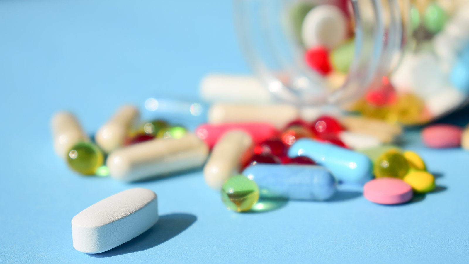 multicolored bright various type pills and capsules spilling out of a toppled white pill bottle. colorful pills and tablets on blue background. pills medicines recall